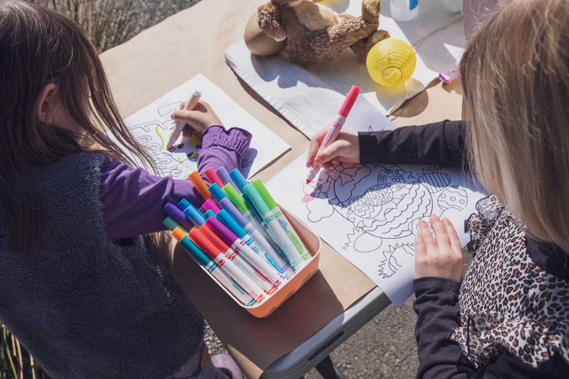 Deux enfants colorient des dessins sur une table, des feutres de toutes les couleurs au premier plan.